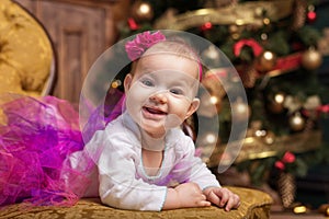 Cute baby girl wearing pink skirt and red headband, laying on couch in front of christmas tree. Smiling toddler.