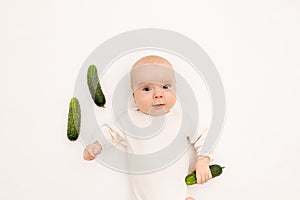 Cute baby girl in white Boda on a white isolated background eating cucumbers, first bait, baby 3-6 months among vegetables, space photo