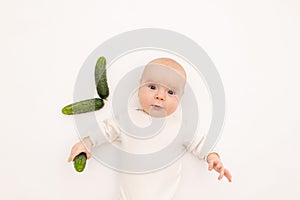 Cute baby girl in white Boda on a white isolated background eating cucumbers, first bait, baby 3-6 months among vegetables, space photo