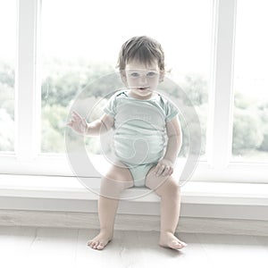 Cute baby at home in white room is sitting near window. The beautiful baby could be a boy or girl and is wearing body suit.