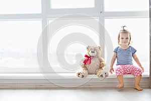 Cute baby at home in white room is sitting near window. The beautiful baby girl with teddy bear.