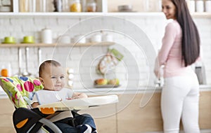 Cute baby playing on high chair in kitchen, mother washing dishes