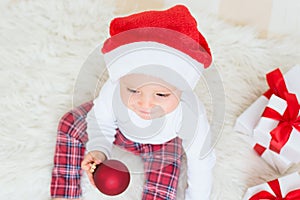 Cute baby with red Santa Claus hat and gift box play with red christmas ball on white bed. Christmas congratulations concept with