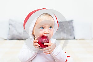Cute baby with red Santa Claus hat play with red christmas ball on white bed. Kid with blue eyes looking upwards. Christmas