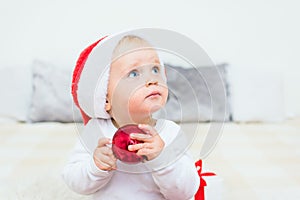 Cute baby with red Santa Claus hat play with red christmas ball on white bed. Kid with blue eyes looking upwards. Christmas