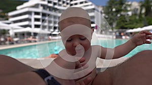 A cute baby sitting and bitting his father`s finger at a swimming pool