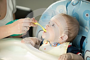 Cute baby sitting in highchair and opening mouth for spoon with