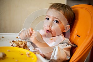 A cute baby is sitting in an orange child seat with a table with dirty hands and face, looking at camera and grimacing