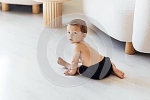 A cute baby is sitting on the white wooden floor in the living room. The little boy toddler is playing at home. The sweet child