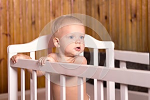 Cute baby standing in a white bed. Nursery for young children in the country house. Little boy learning to stand in his crib.