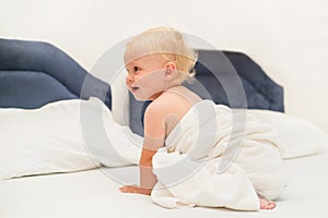 Cute baby under a white towel sits on the bed