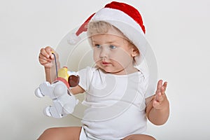 Cute baby wearing christmas hat and body suit playing with plastic dog, charming infant holding his toy, child looking away, baby