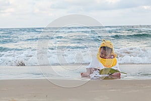 Cute baby wearing a yellow cartoon bathrobe sitting and playing on the beach near the sea