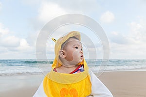 Cute baby wearing a yellow duck cartoon bathrobe sitting and playing on the beach near the sea