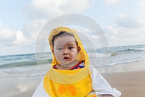 Cute baby wearing a yellow duck cartoon bathrobe sitting and playing on the beach near the sea