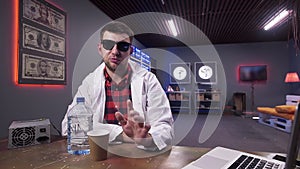 Cute bearded guy in white coat sits behind desk with plastic bottle and cup