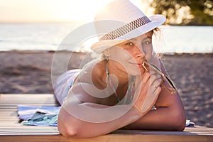 Cute beautiful attractive tanned lady in hat lies on deckchair on sea beach at sunset on warm summer evening. Pretty woman smiles