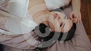 Cute beautiful caucasian brunette bride in white peignoir and lingerie with beige manicure lies in bed with hand on her