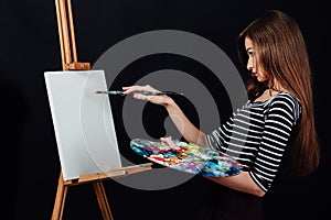 Cute beautiful girl artist painting a picture on canvas an easel. Space for text. Studio black background.