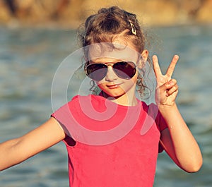 Cute beautiful kid girl in fashion sunglasses showing v sign making the selfie on blue sea and sky background. Closeup toned port