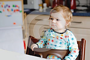 Cute beautiful little baby girl at home or nursery, indoors. Happy healthy toddler child sitting by the table.