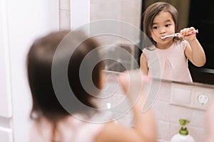 Cute beautiful little girl is brushing her teeth with tooth brush in front of the bathroom mirror