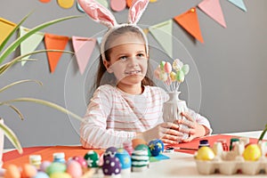 Cute beautiful little girl wearing rabbit ears sitting at table preparing for Easter against gray wall with decorations, kid