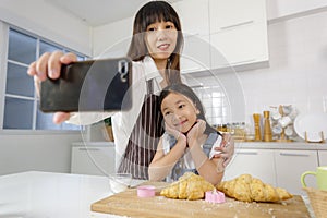 A cute and beautiful mother with young and little daughter, 7 years old, taking a selfie photo with a smartphone in modern kitchen