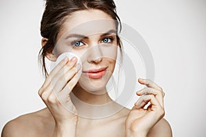 Cute beautiful natural brunette girl cleaning face with cotton sponge smiling looking at camera over white background