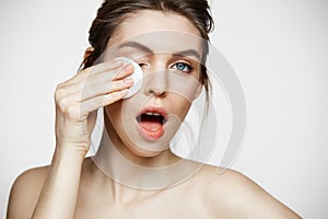 Cute beautiful natural brunette girl cleaning face with cotton sponge smiling looking at camera over white background