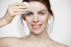 Cute beautiful natural brunette girl cleaning face with cotton sponge smiling looking at camera over white background