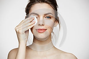 Cute beautiful natural brunette girl cleaning face with cotton sponge smiling looking at camera over white background