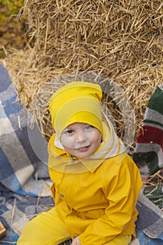 Cute beautiful prescholer boy in an orange pants, raincoat, hat, rubber boots near a pile of hay. Cosiness, autumn