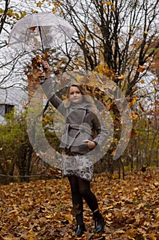 Cute beautiful teenage brunette girl in an grey coat holding a transparent umbrella with falling leafes over her. Cosiness, autumn