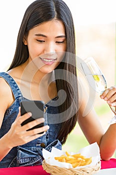 A cute beautiful young Asian woman sitting alone at the fast food shop and holding wine glass and smartphone