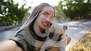 A cute beige pug puppy is trying to lick the face. A woman holds camera and squints