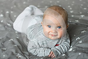 Cute blond baby with blue eyes is laying on grey bed view from above