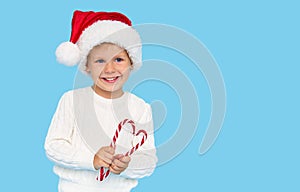 A cute blond baby boy in a Santa Claus hat holding a heart-shaped lollipop cane on a blue background.