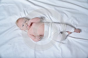 Cute blond baby with fingers in his mouth lies on a white sheet. Top view