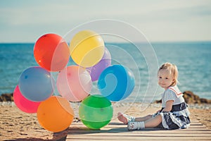 Cute blond baby girl child posing enjoying summer life time on sandy beach sea side on wooden pier with colourful plenty balloons