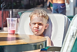 Cute blond boy sits at table drinking milkshake on vacation