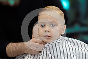 Cute blond caucasian baby boy with blue eyes in a barber shop after having haircut by hairdresser. Hands of stylist. Children fash photo