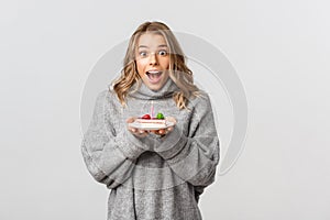 Cute blond girl celebrating birthday, holding b-day cake and smiling happy, standing over white background
