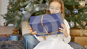 Cute blond girl has recieved a gift box wrapped in craft dark blue paper sitting near christmas tree, happy childhood