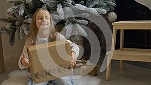 Cute blond girl has recieved a gift box wrapped in craft paper sitting near christmas tree, happy childhood and xmas