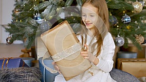 Cute blond girl has recieved a gift box wrapped in craft paper sitting near christmas tree, happy childhood and xmas