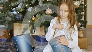 Cute blond girl is opening dark blue gift box she recieved sitting near christmas tree, happy childhood and xmas