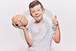 Cute blond kid holding brain smiling happy and positive, thumb up doing excellent and approval sign