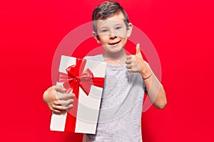 Cute blond kid holding gift smiling happy and positive, thumb up doing excellent and approval sign