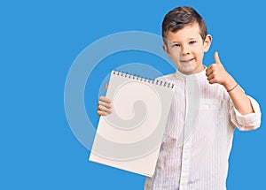 Cute blond kid holding notebook smiling happy and positive, thumb up doing excellent and approval sign